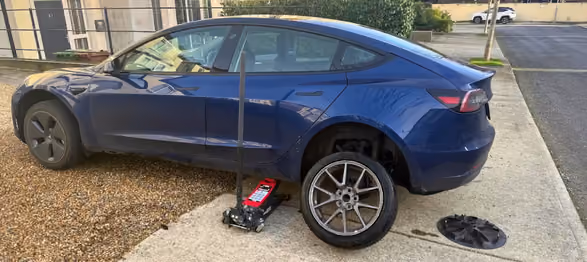 Blue car lifted by jack with rear wheel removed and tire leaning against car on driveway.