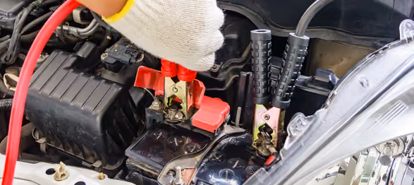 Gloved hand connecting jumper cables to a car battery in an engine compartment.