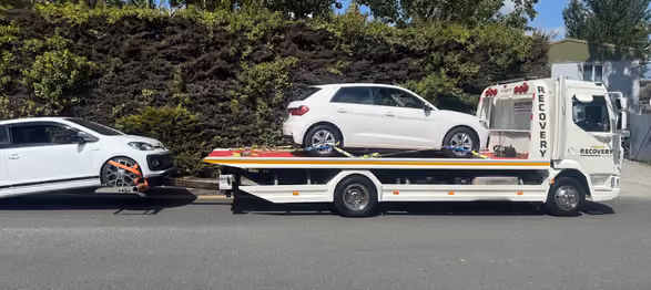 White tow truck carrying a white car and towing another white car on a road with trees behind.