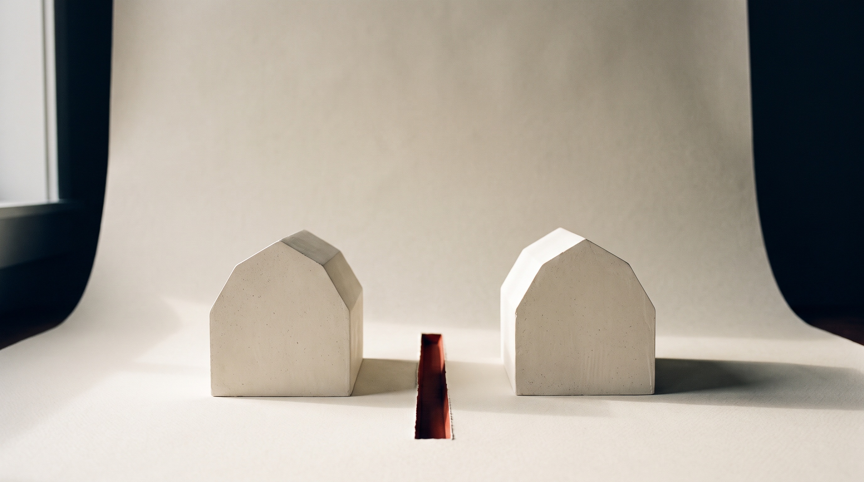 Two identical plaster models of the same building stand on a bone-white surface, divided by a single narrow scarlet line cut into the ground between them.