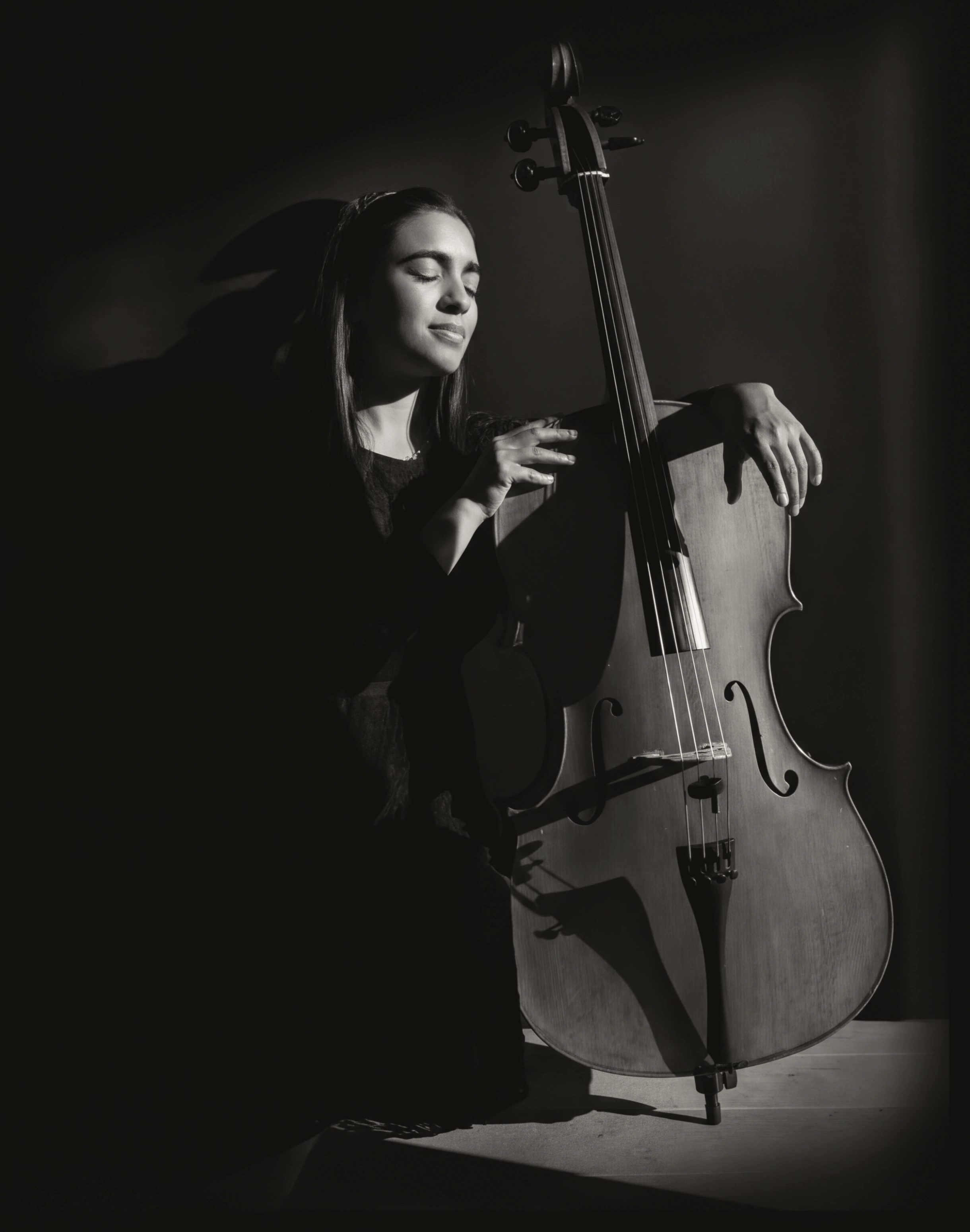 Woman with eyes closed leaning on a cello, shown in black and white with dramatic lighting.