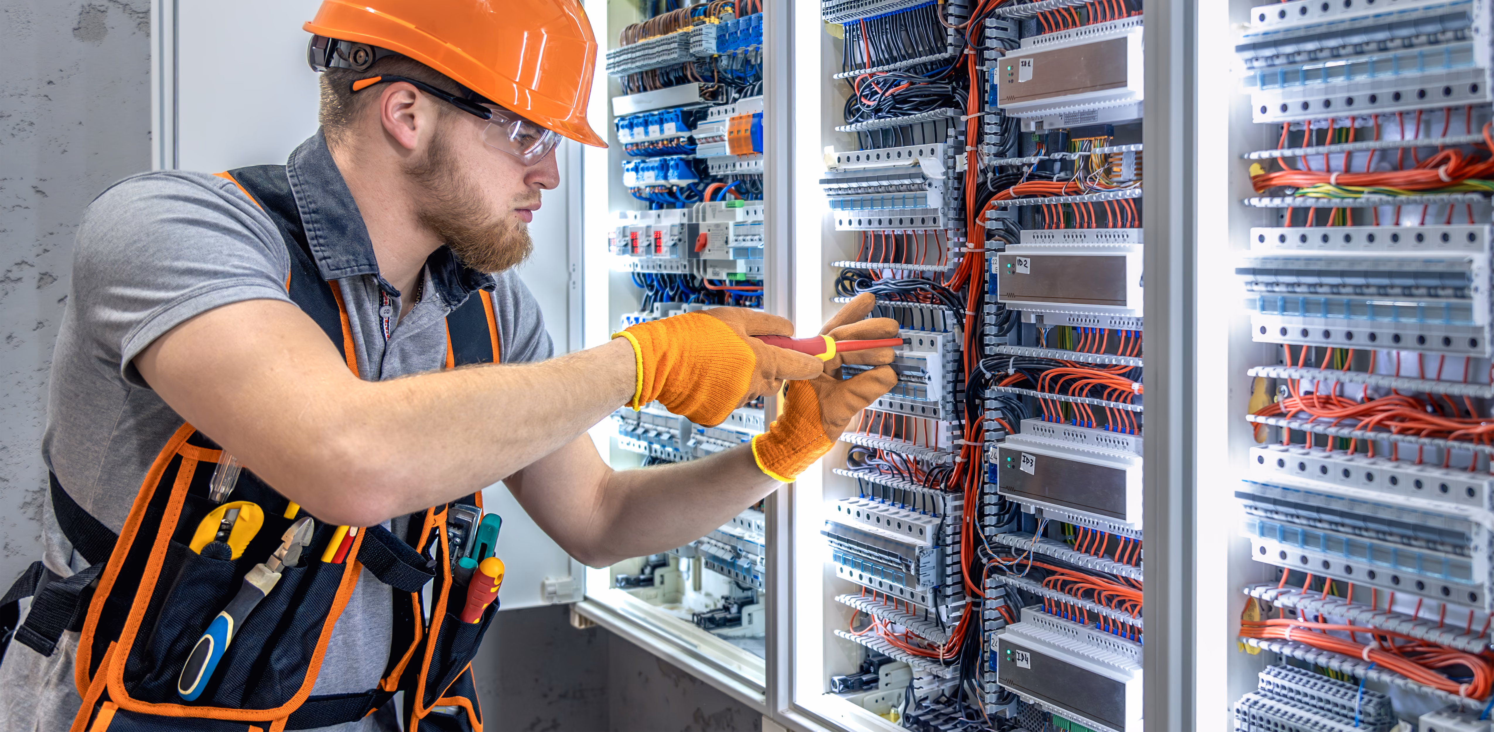 Hombre con casco y guantes naranjas trabaja en un panel eléctrico lleno de cables y componentes.
