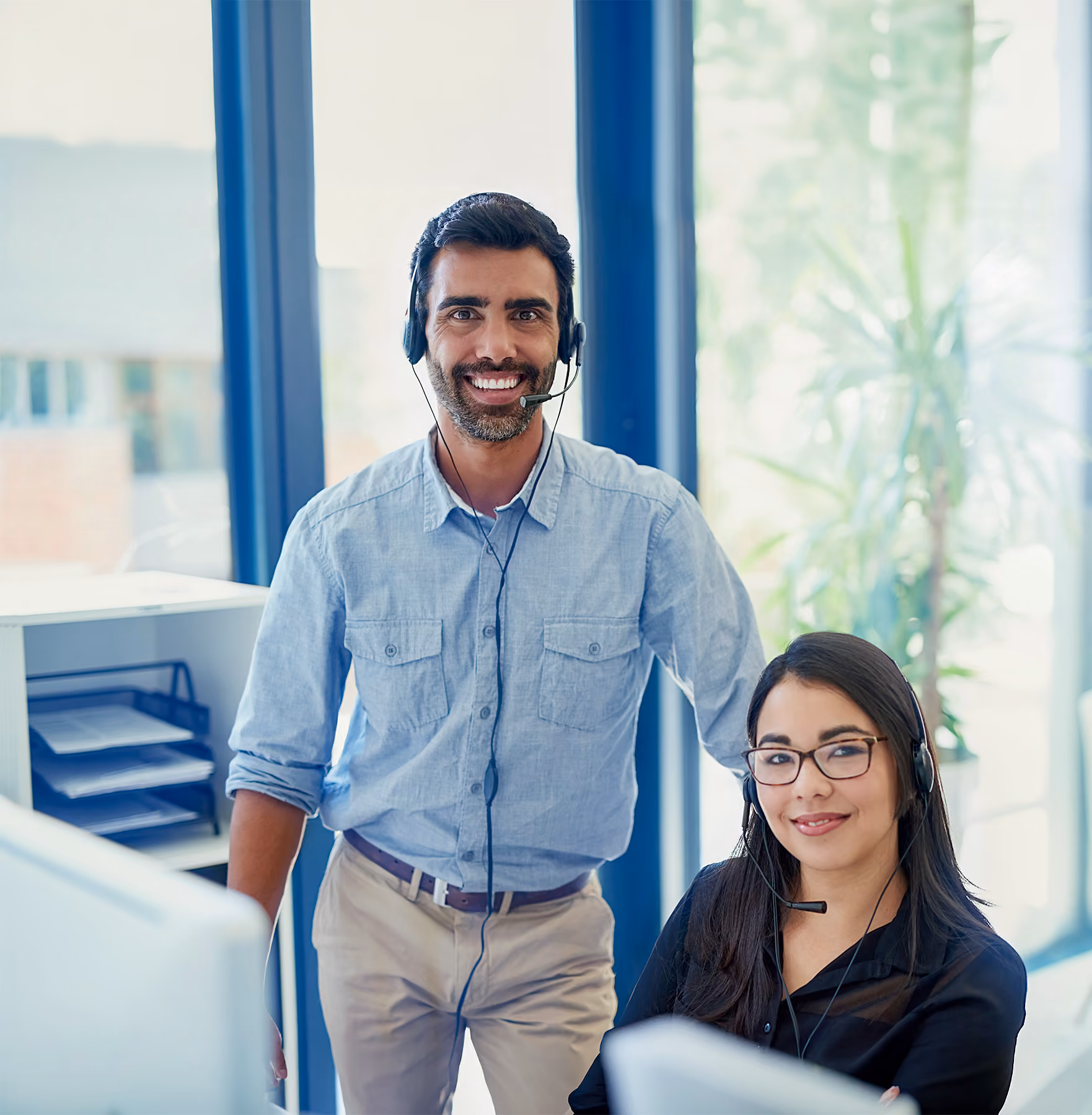 Hombre y mujer con auriculares y micrófono sonríen en oficina luminosa de servicio al cliente.