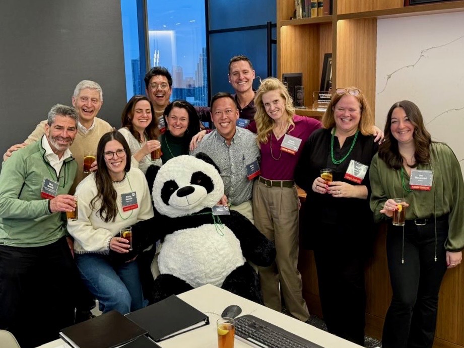 Group of people smiling and holding drinks posing around a large stuffed panda in an office.