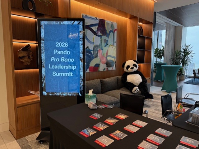 Event sign reading '2026 Pando Pro Bono Leadership Summit' beside table and a large panda plush on a couch.