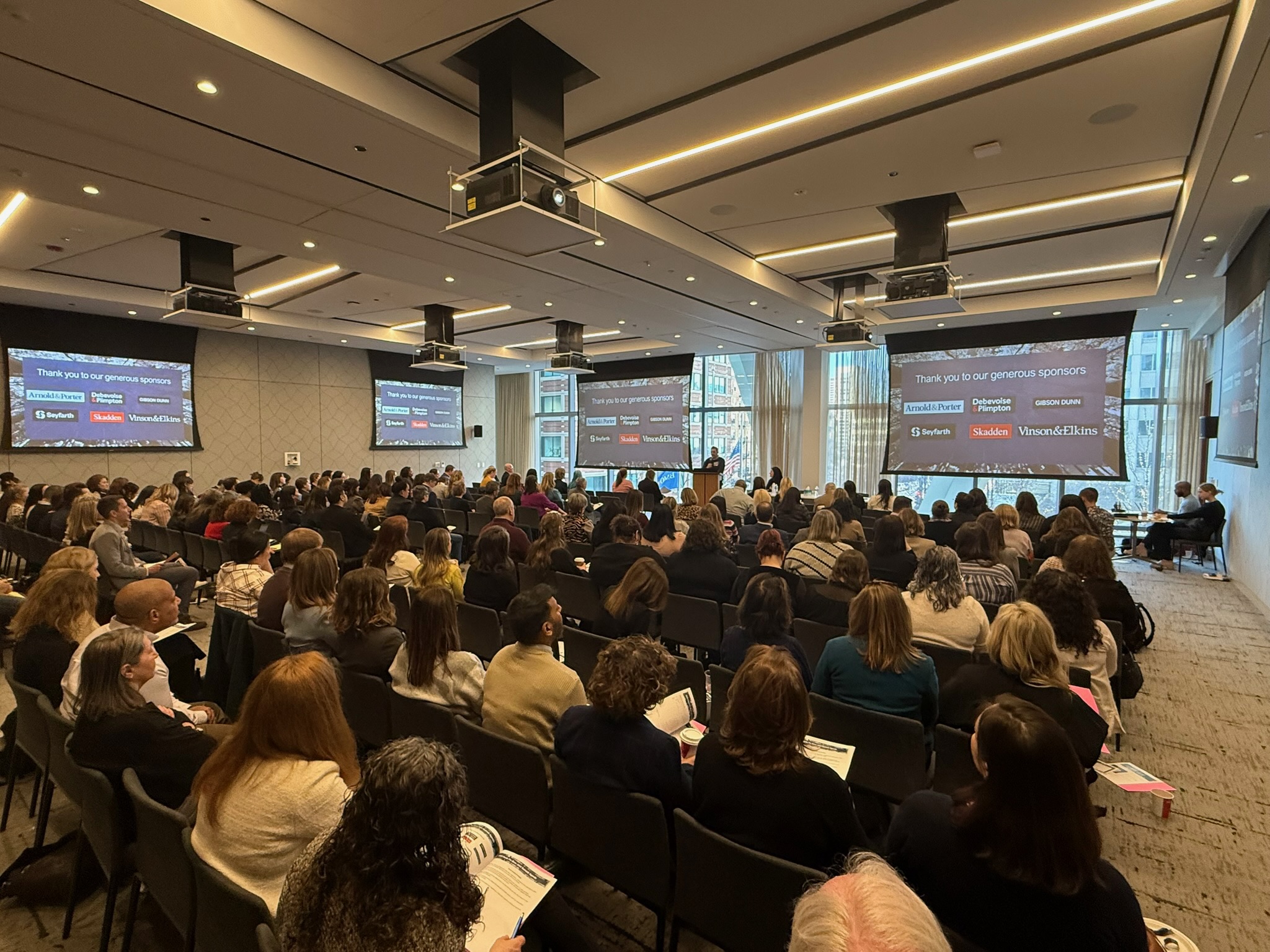 Audience seated in a conference room watching a speaker and multiple screens showing sponsor logos.