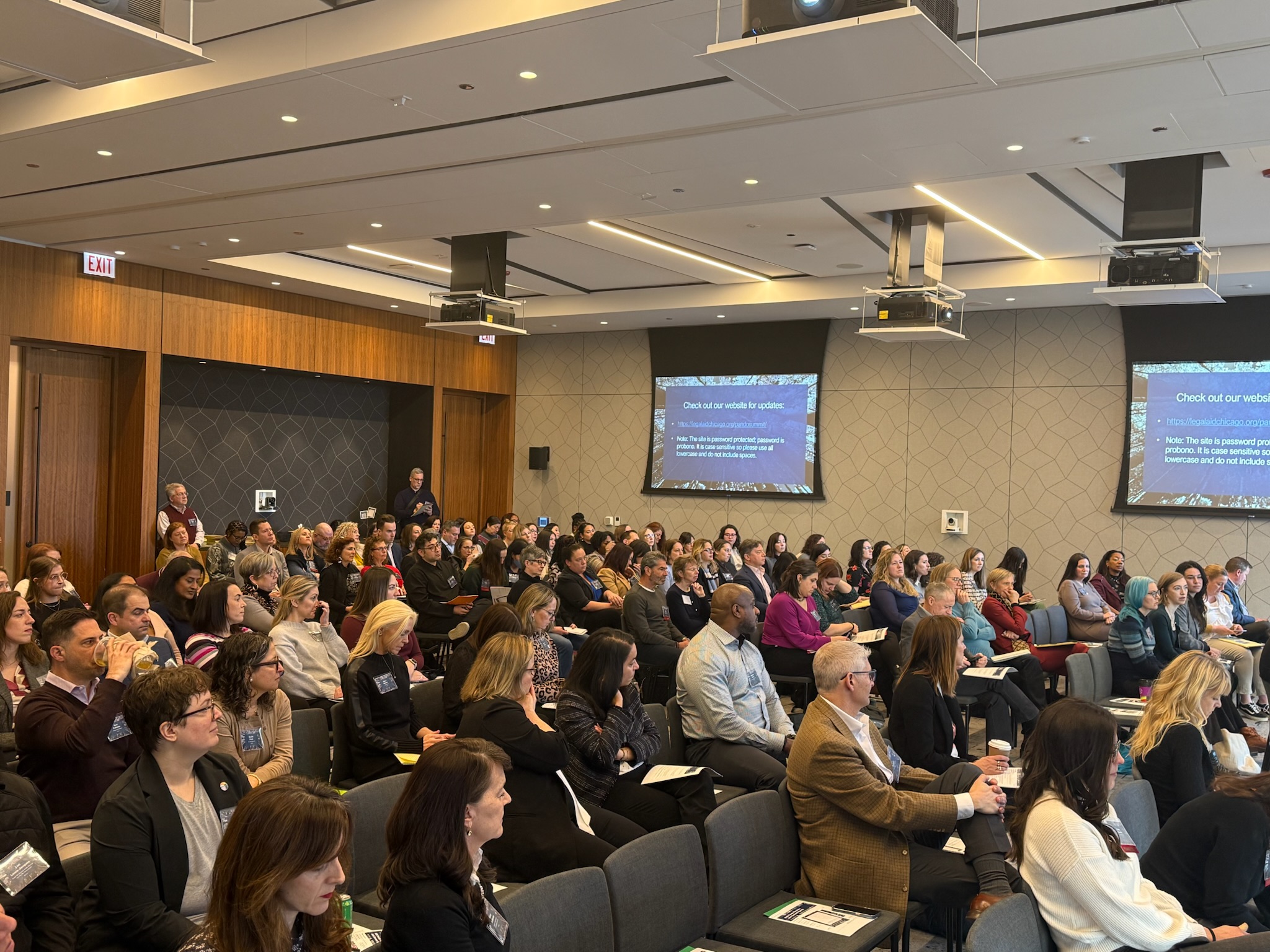 Audience of diverse adults seated attentively in a conference room with two screens showing update info.
