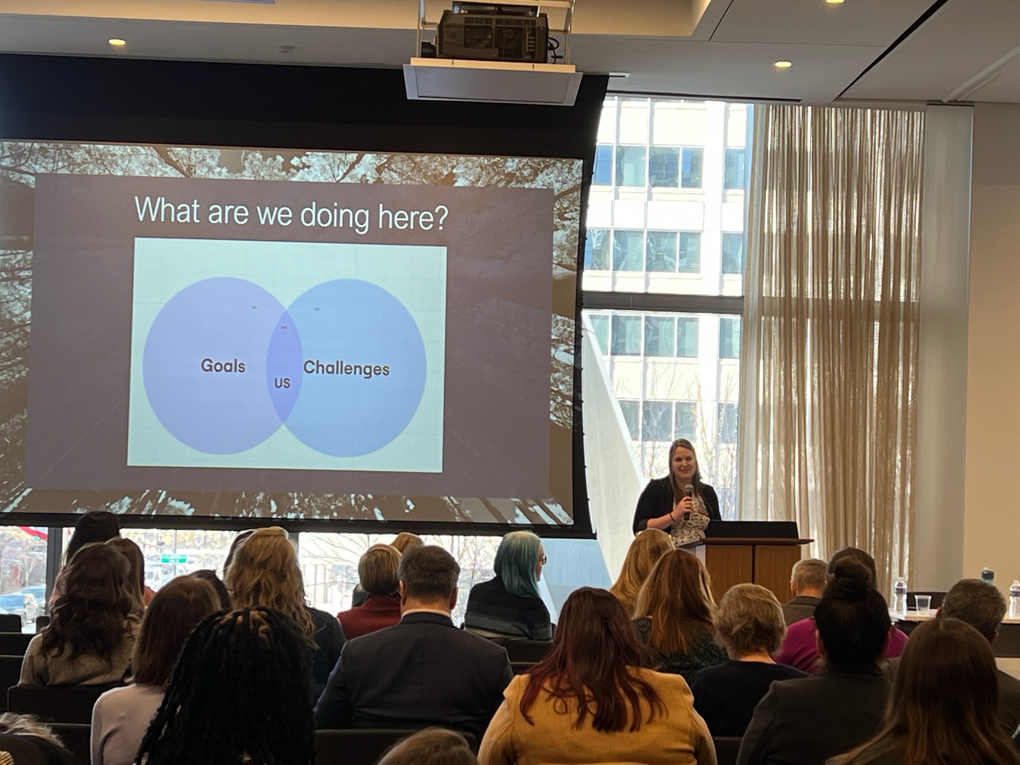 Audience watching a speaker near a large screen showing a Venn diagram titled 'What are we doing here?'