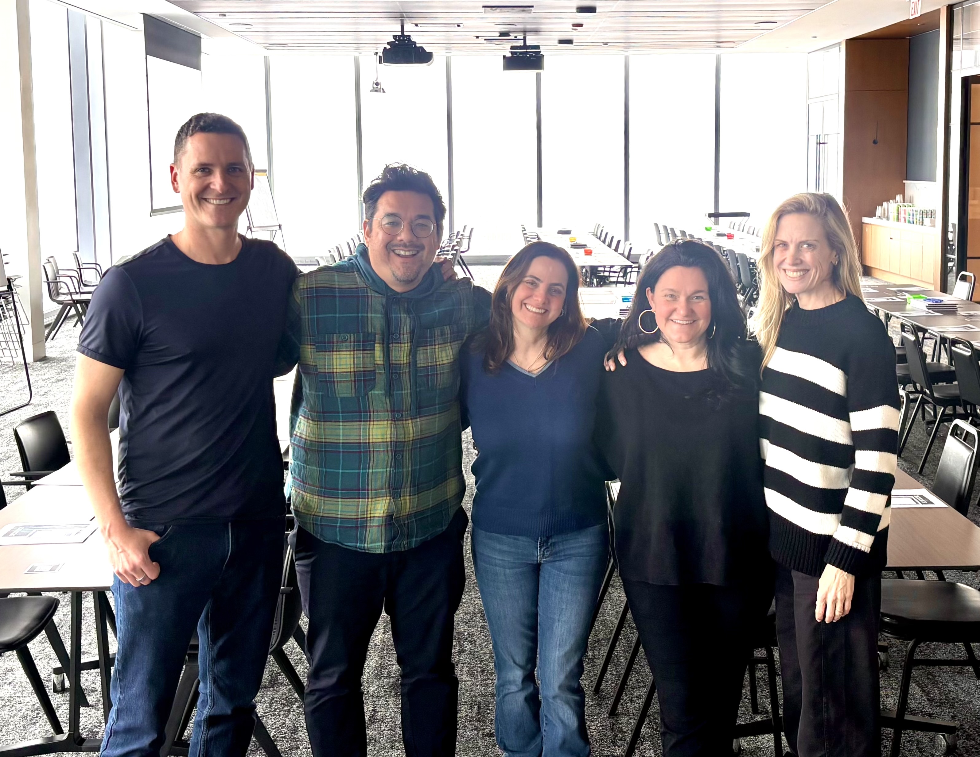 Five smiling adults standing arm-in-arm in a well-lit conference room with rows of chairs and tables.