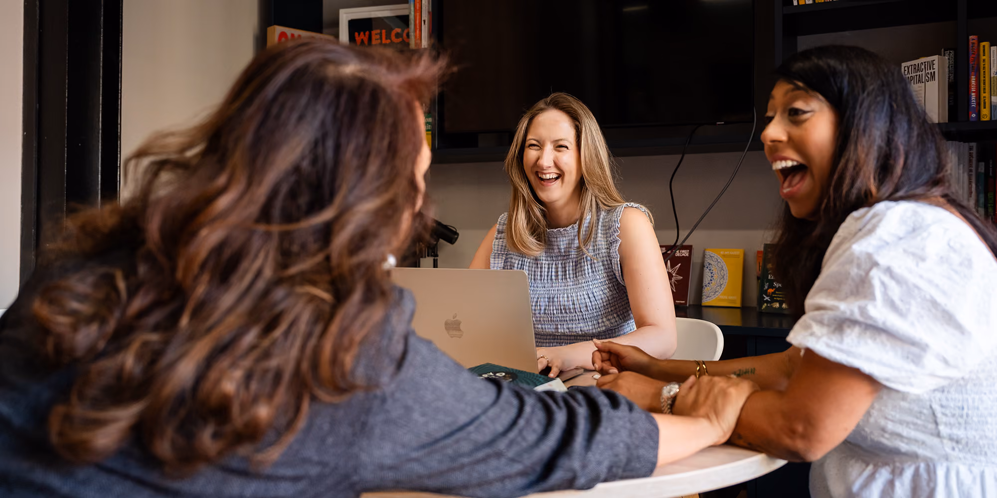 Three women sitting around a table, smiling and laughing during a meeting with a laptop in front.