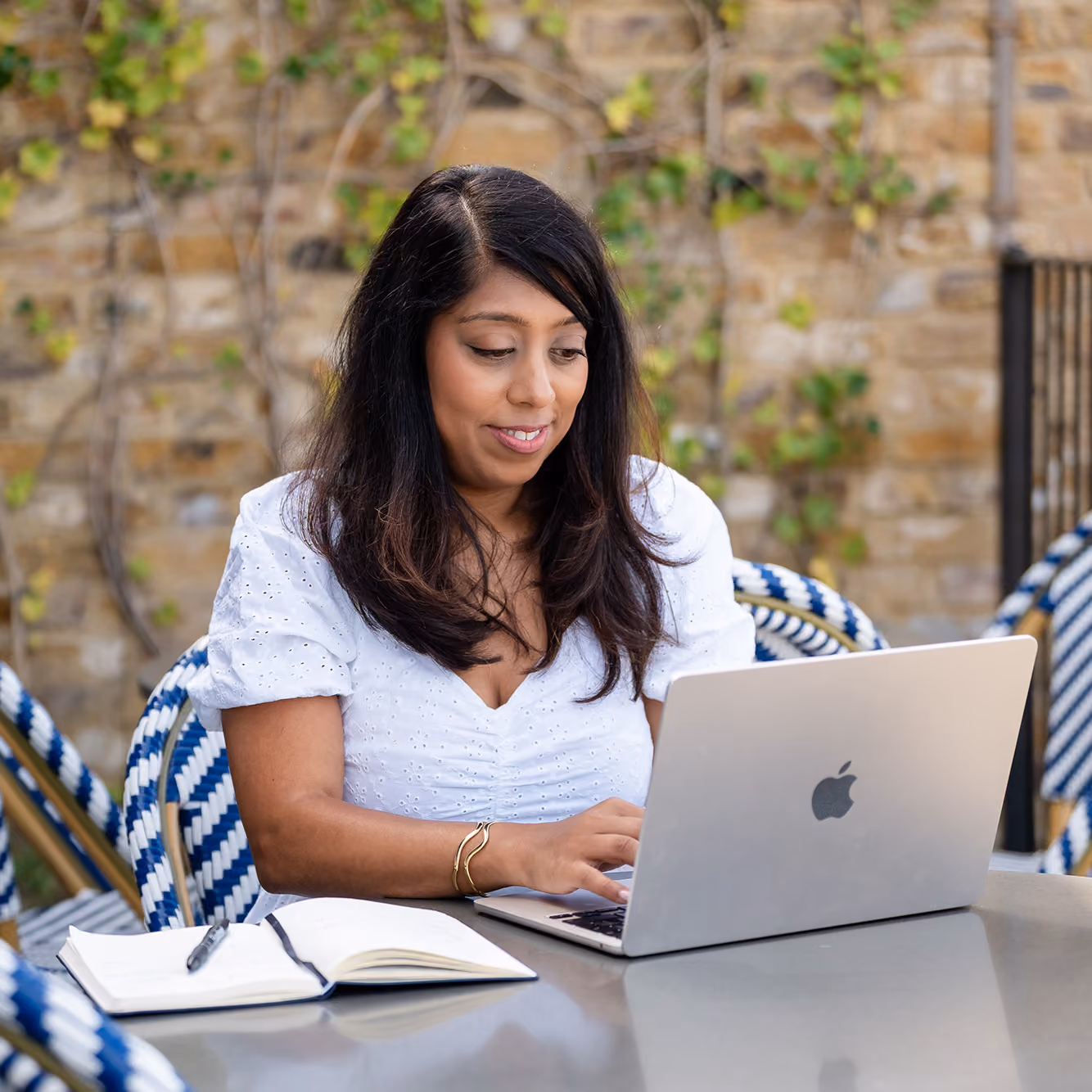 Woman in a white blouse typing on a laptop at an outdoor cafe table with a notebook and pen.