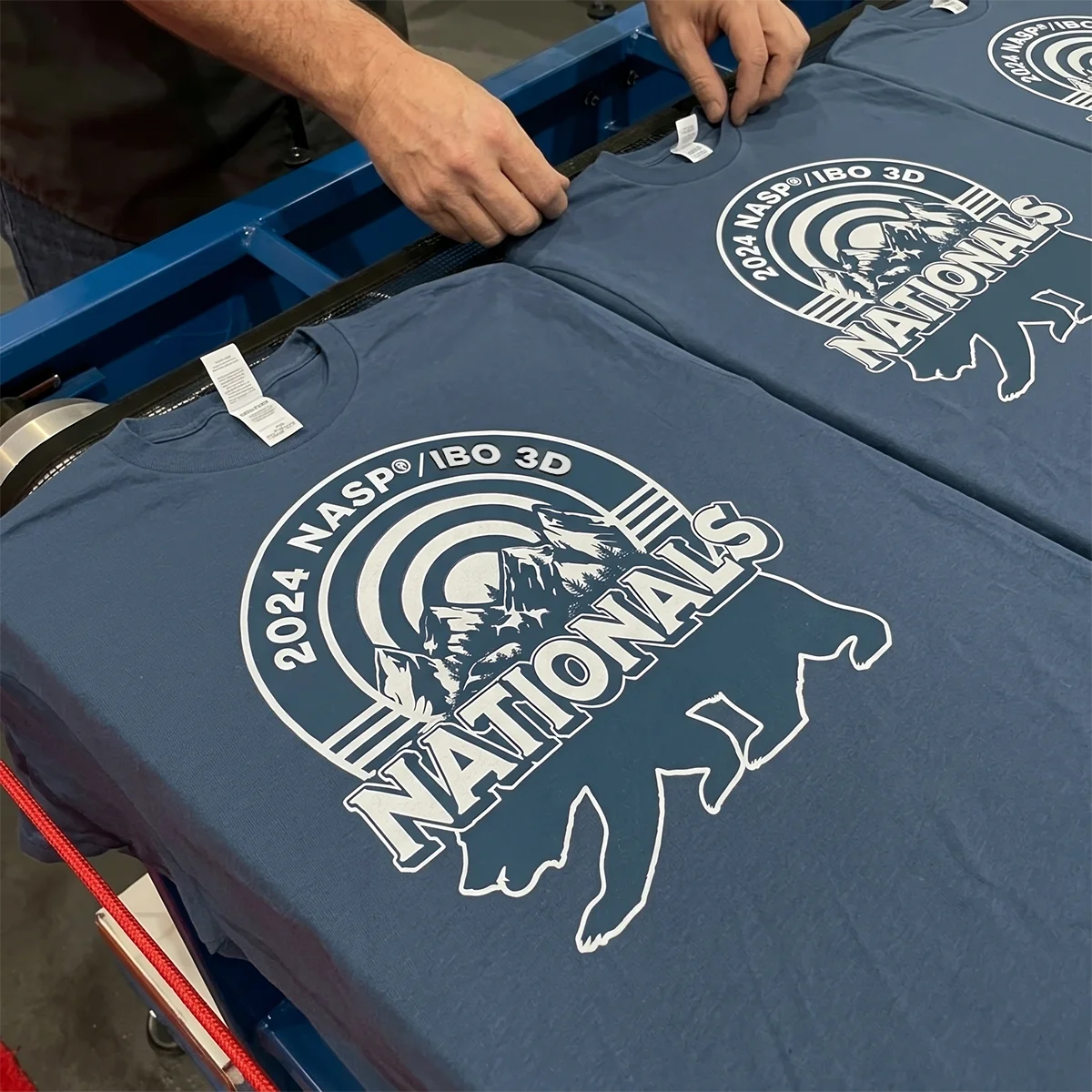 Hands arranging blue T-shirts printed with 2024 NASP IBO 3D Nationals logo showing mountains and bear.