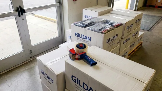 Stack of Gildan boxes with a tape dispenser on top in a warehouse near glass doors.