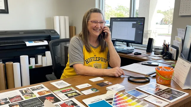 Smiling woman in a Hufflepuff shirt talks on phone at desk with color swatches and printed materials.
