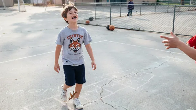 Happy boy wearing a gray Coyotes t-shirt standing on a cracked playground with basketballs nearby.
