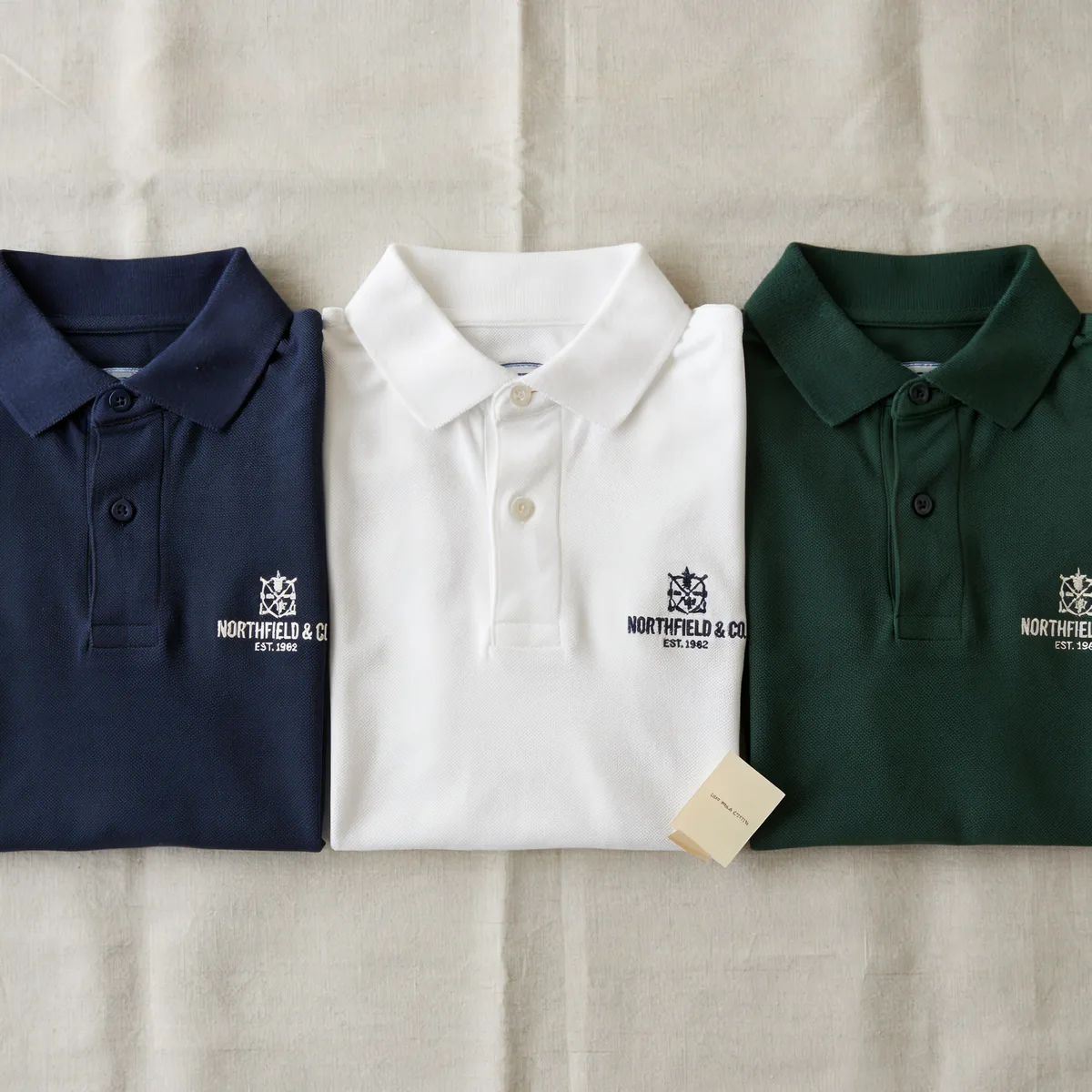 Three folded polo shirts in navy, white, and green with Northfield & Co logos.