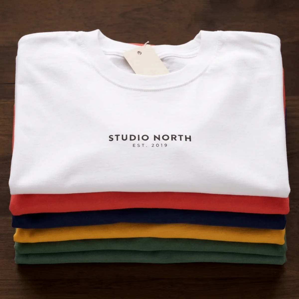 Stack of folded t-shirts topped by a white tee with 'STUDIO NORTH EST. 2019' printed on it.