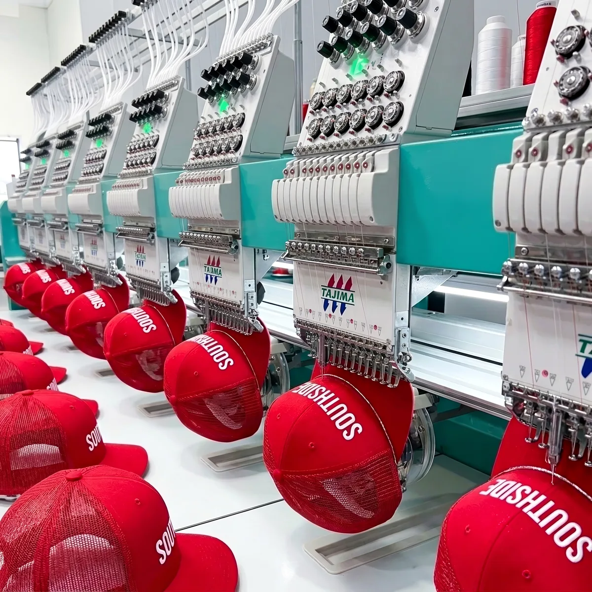 Row of industrial embroidery machines stitching white text on red trucker hats in a factory.