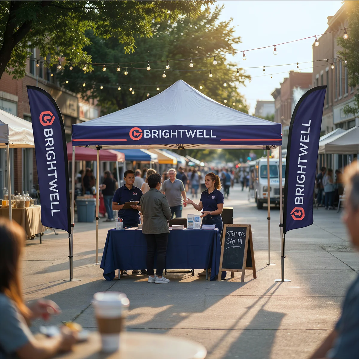 Brightwell booth with staff talking to a visitor at an outdoor street event with string lights above.