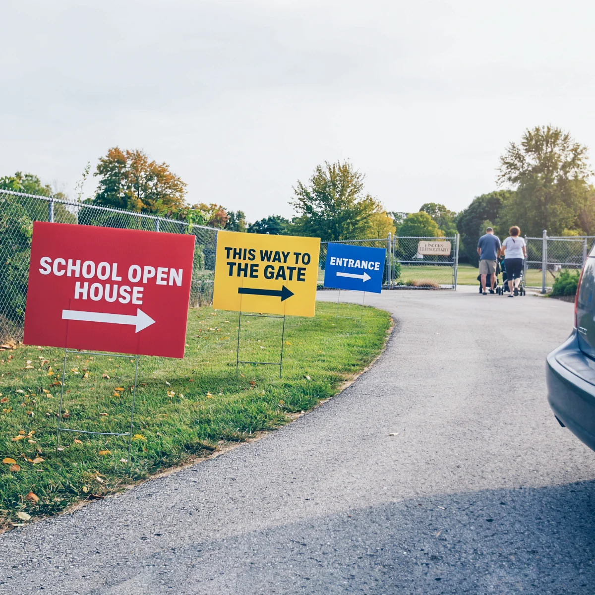 Signs on grass directing to a school open house, gate, and entrance with people walking in the background.