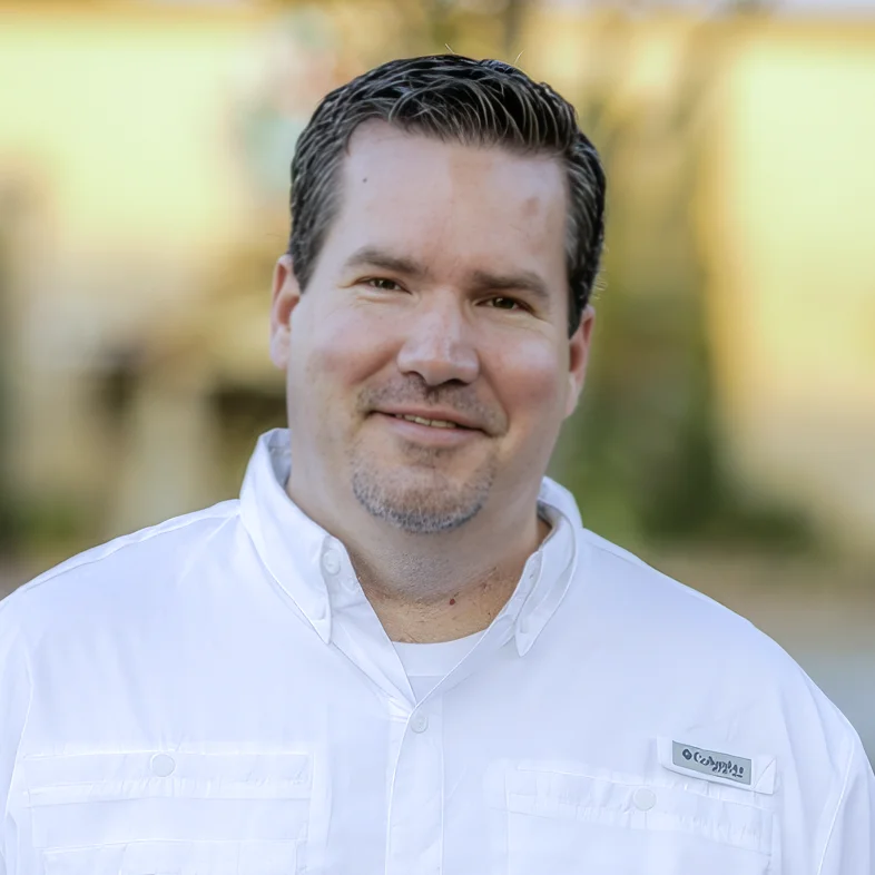 Man with short dark hair, goatee, and a white button-up shirt smiling outdoors with blurred background.