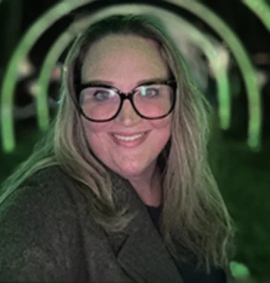 Smiling woman with long hair and large glasses under green arched lights at night.