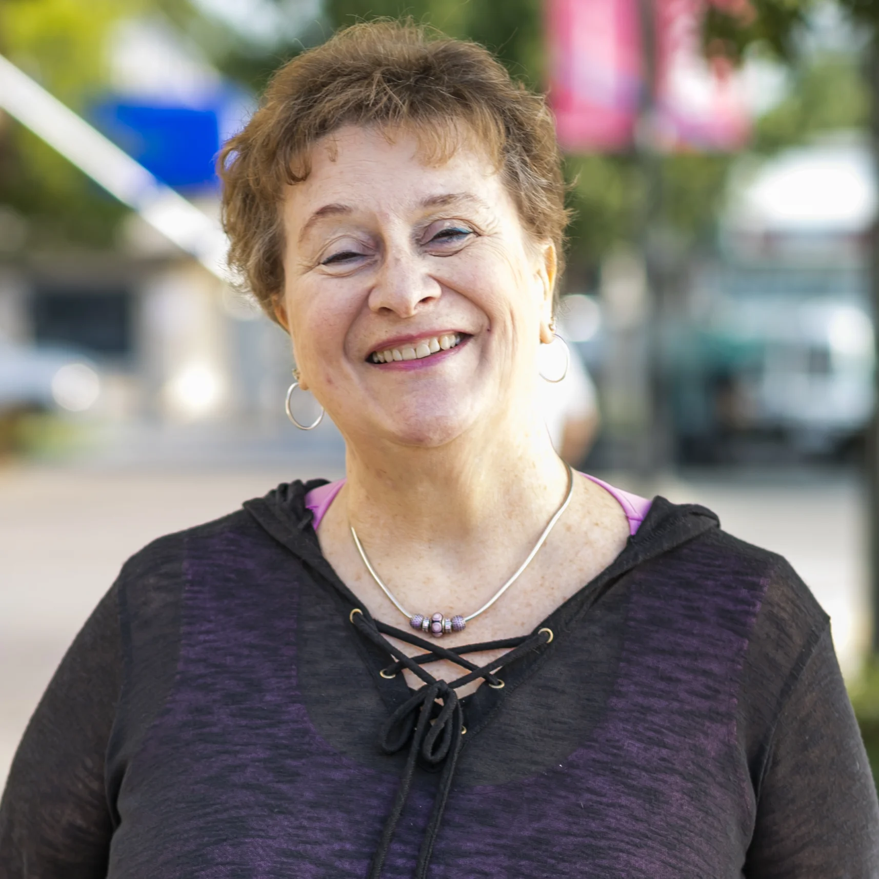 Smiling older woman with short brown hair wearing hoop earrings, necklace, and a dark lace-up top outdoors.