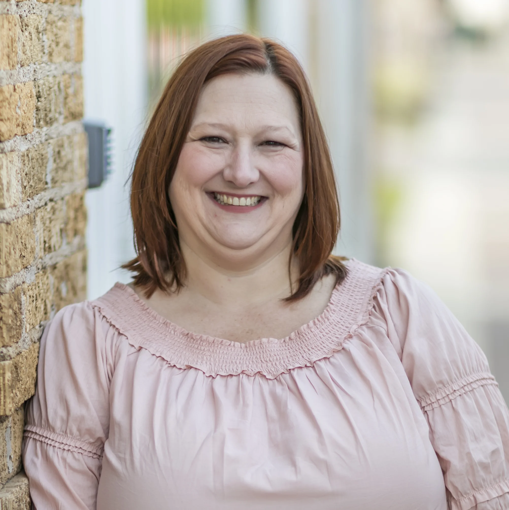 Smiling woman with shoulder-length red hair wearing a pink blouse leaning against a brick wall.