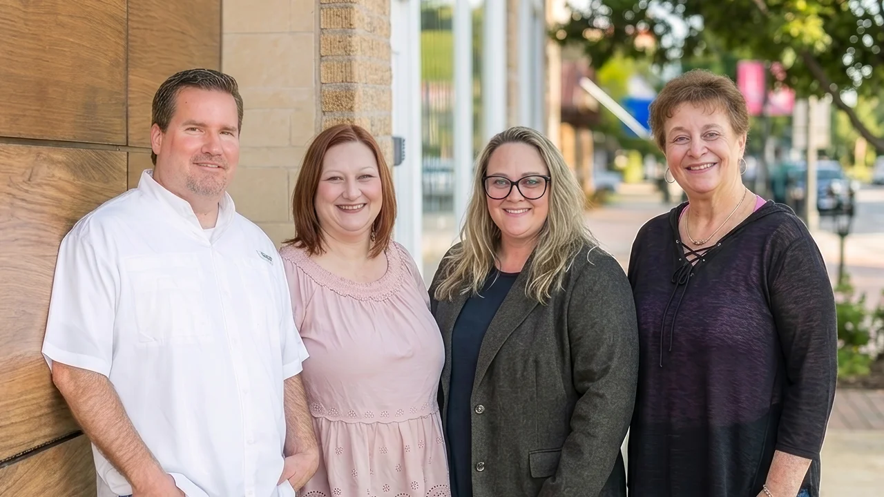 Four adults standing outdoors smiling, with a wood wall and street background.
