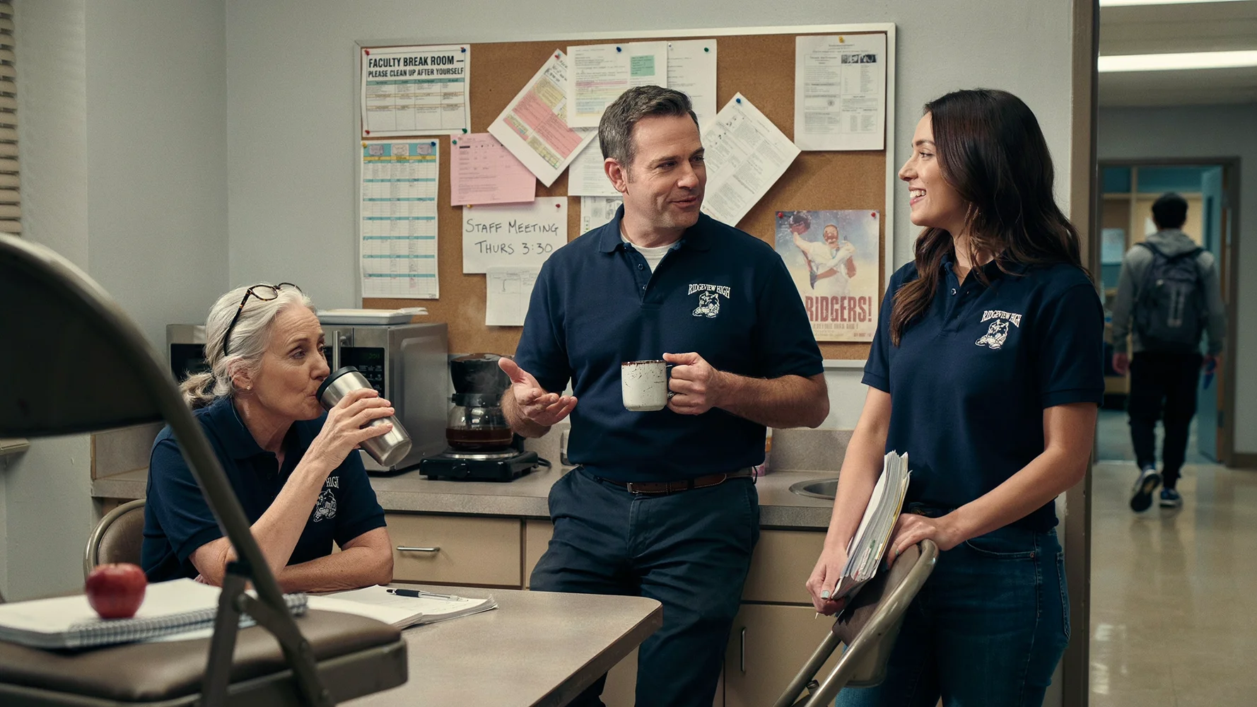 Three teachers in navy Ridgeview High polo shirts chatting in a faculty break room with coffee cups.