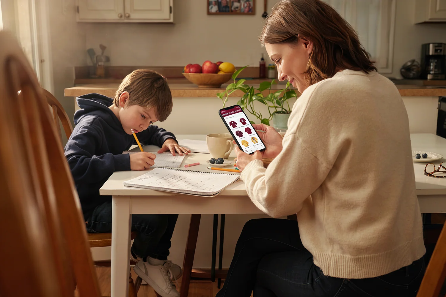 Young boy focused on homework while woman shops for sweaters on a smartphone at the kitchen table.