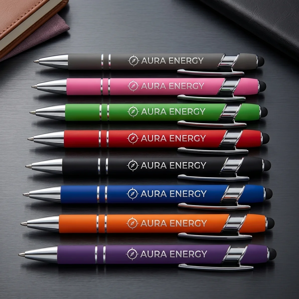 Seven colorful pens with Aura Energy logo arranged horizontally on a dark surface with notebooks nearby.