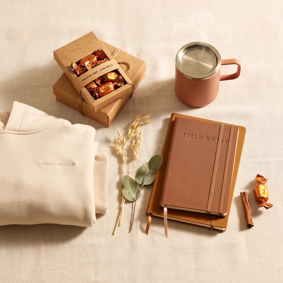 Cozy beige sweatshirt, caramel candies, steaming mug, two notebooks, cinnamon stick, and dried plants on fabric.