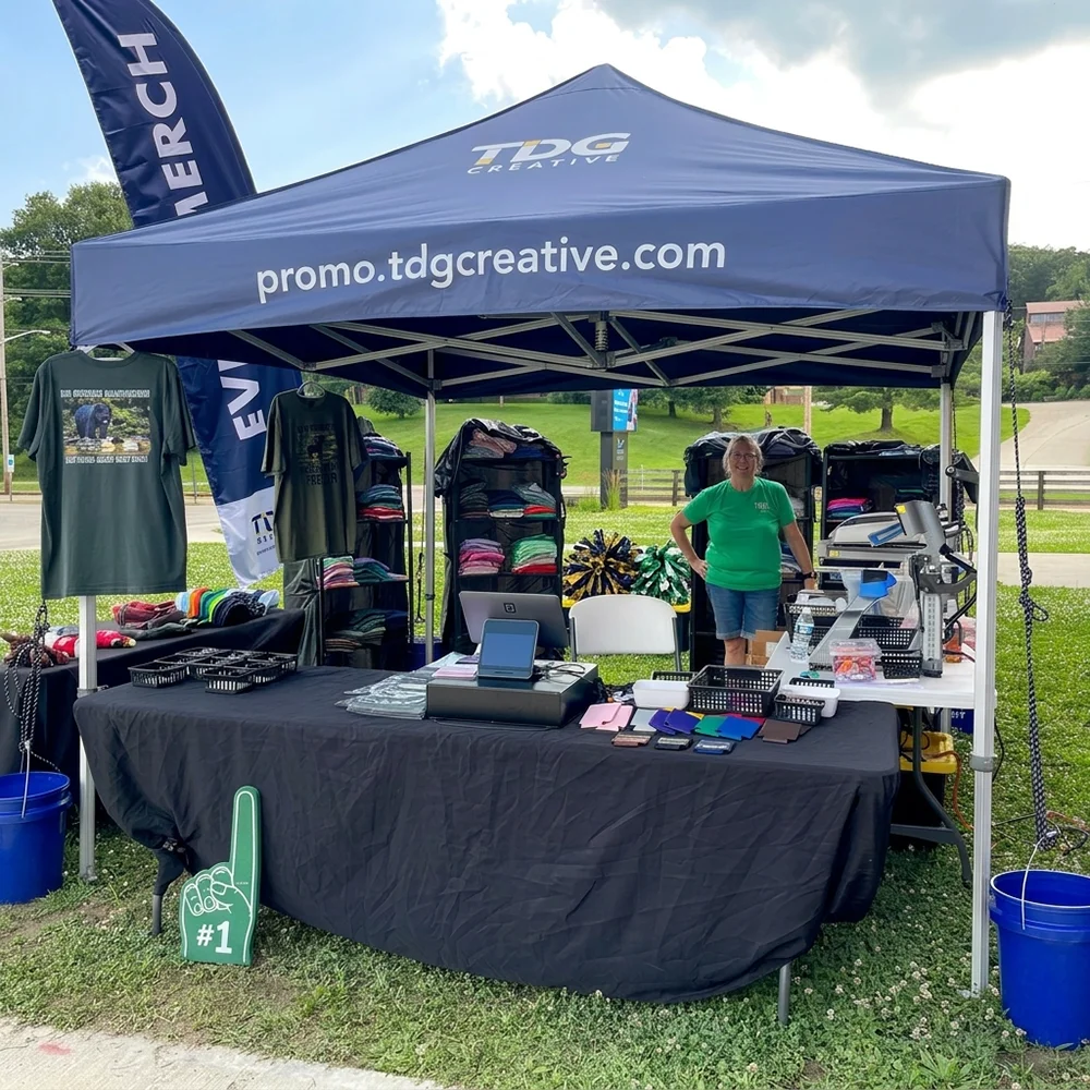 Outdoor booth with TDG Creative tent selling shirts, with woman in green shirt standing inside.