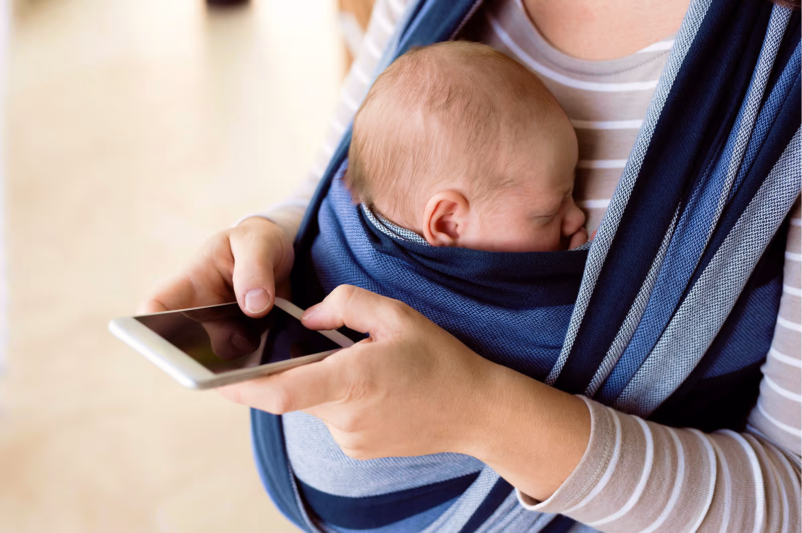 A parent with their baby in a sling, looking at a phone.