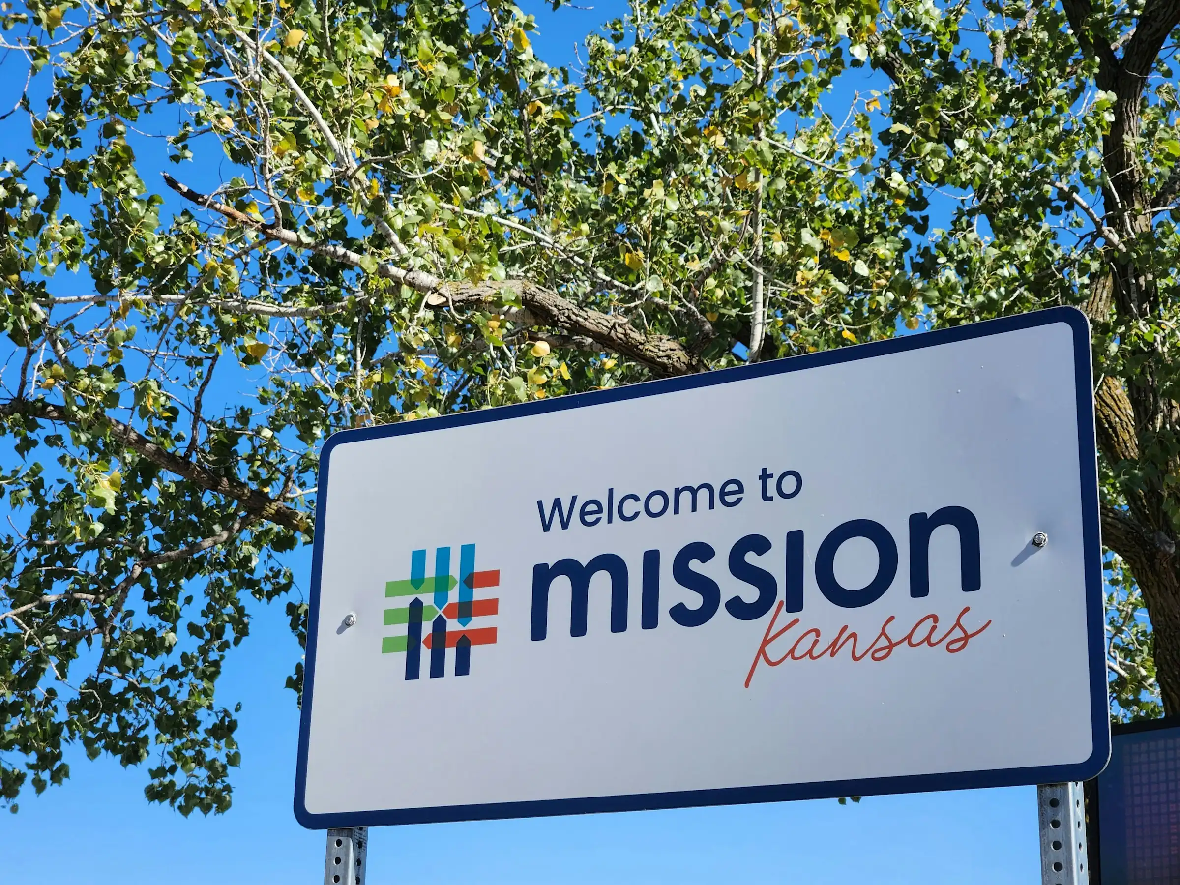 "Welcome to Mission, Kansas" sign against a cottonwood tree and clear blue sky