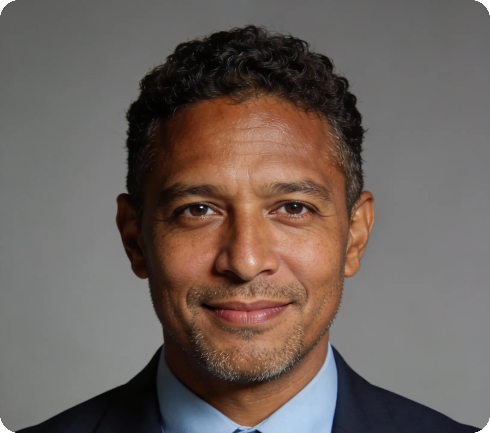 Smiling middle-aged man with curly hair and light stubble wearing a dark suit and blue shirt against a plain gray background.