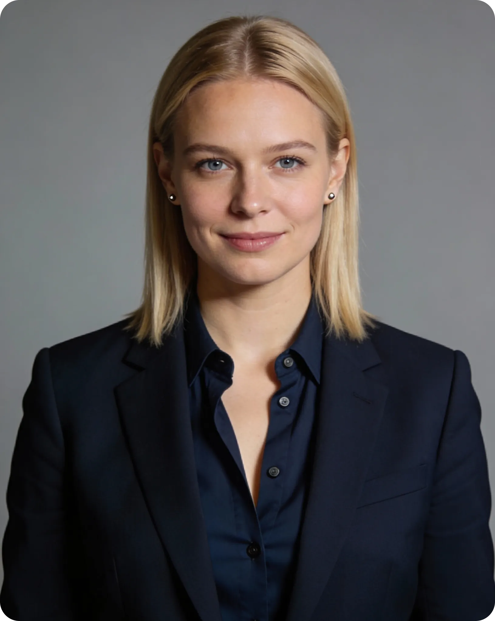Professional portrait of a blonde woman with blue eyes wearing a dark navy blazer and shirt, smiling slightly against a plain gray background.