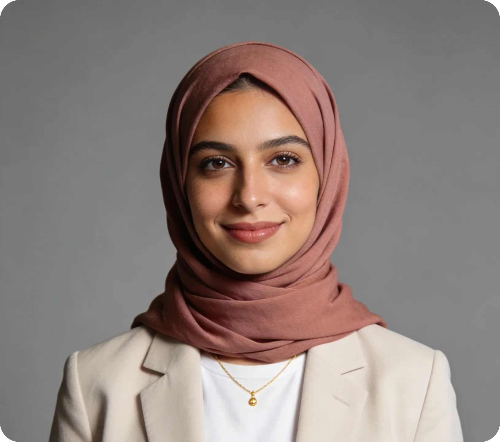 Smiling woman wearing a dusty rose hijab, beige blazer, white top, and gold necklace with a round pendant.