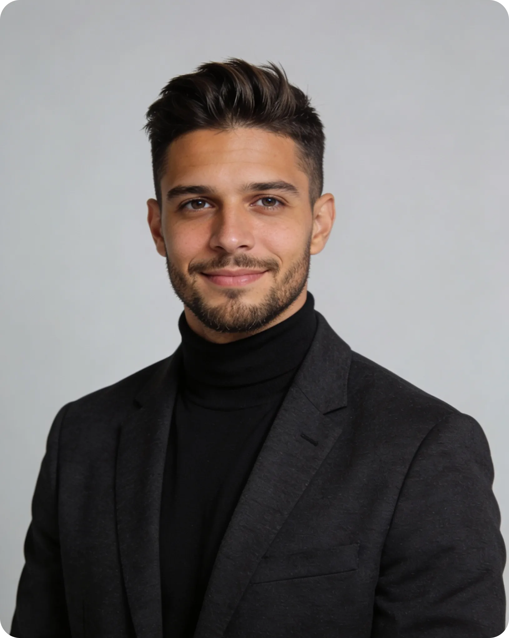 Smiling young man with dark hair and beard wearing a black turtleneck and blazer against a plain gray background.