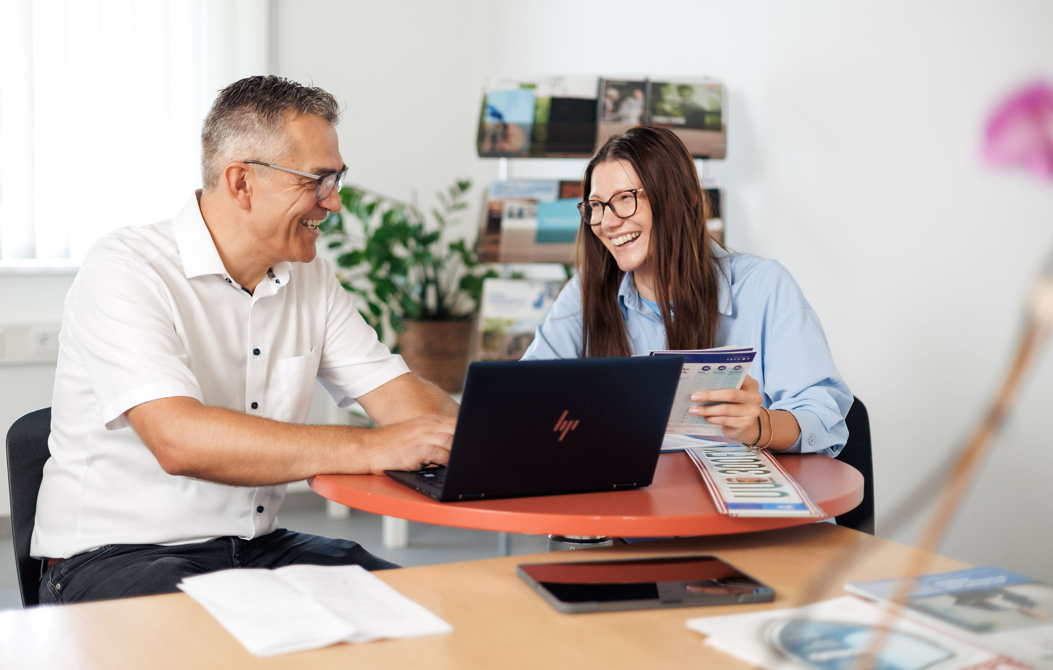 Ein Mann und eine Frau sitzen am Schreibtisch mit einem Laptop und lächeln sich an