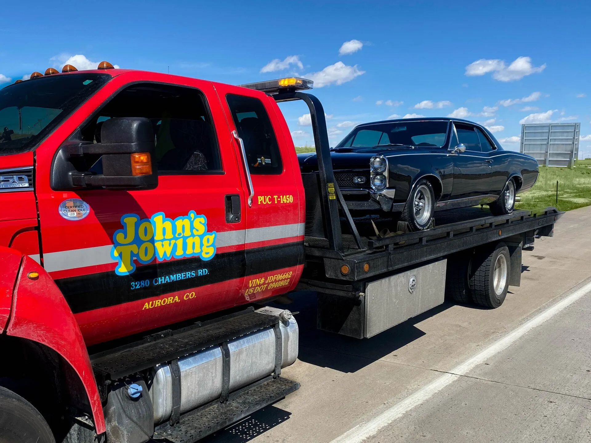 Red John's Towing truck carrying a black classic car on its flatbed in daytime on a highway.