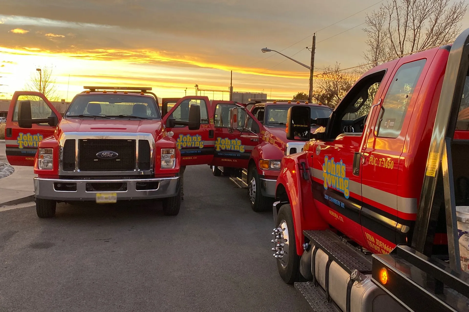 Three red John's Towing trucks with open doors parked on a street at sunset.