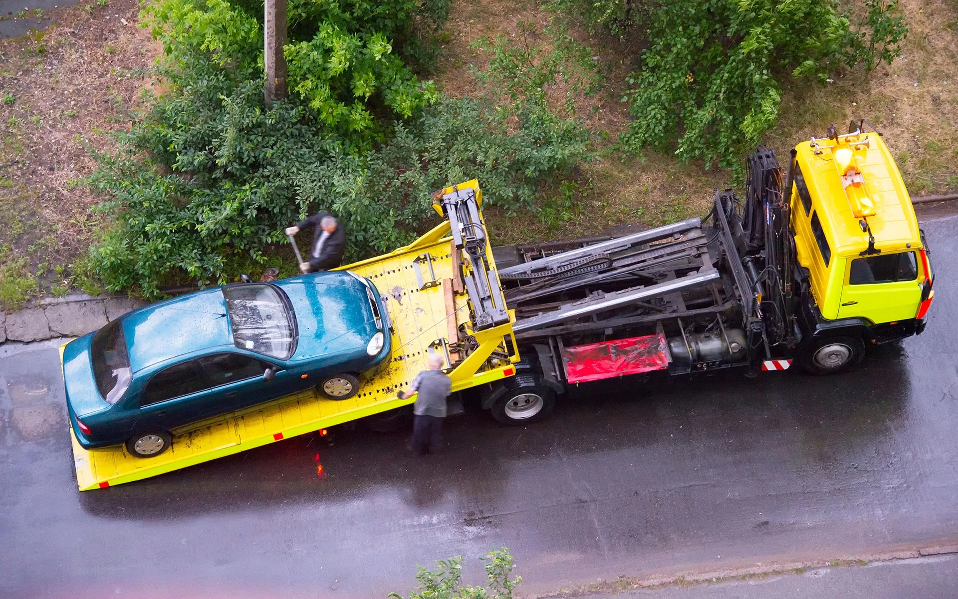 A teal car is being loaded onto a yellow tow truck on a wet street with two men working nearby.