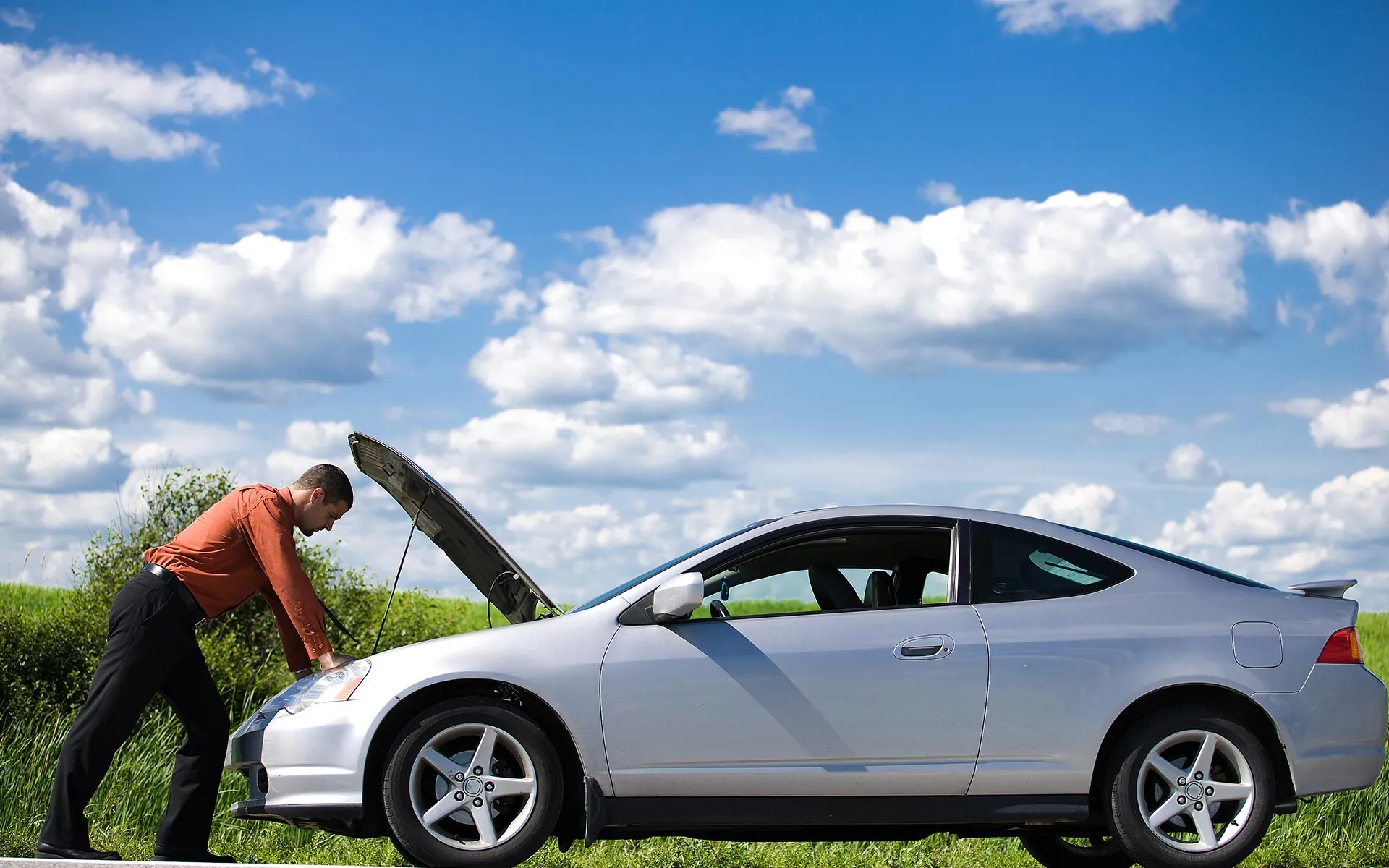 Man in orange shirt inspecting engine of silver car with hood open on roadside.