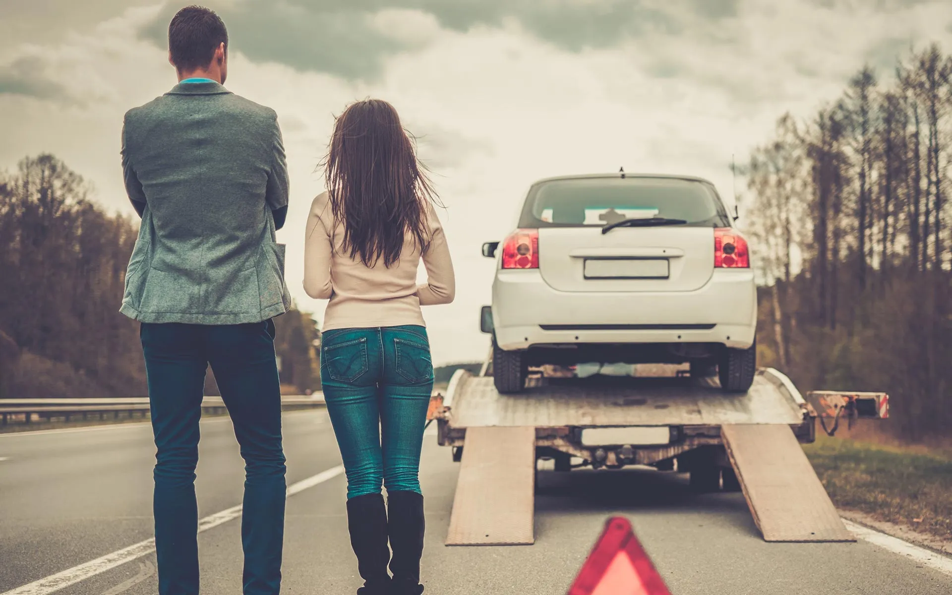 Man and woman standing on roadside watching a white car being loaded onto a tow truck.
