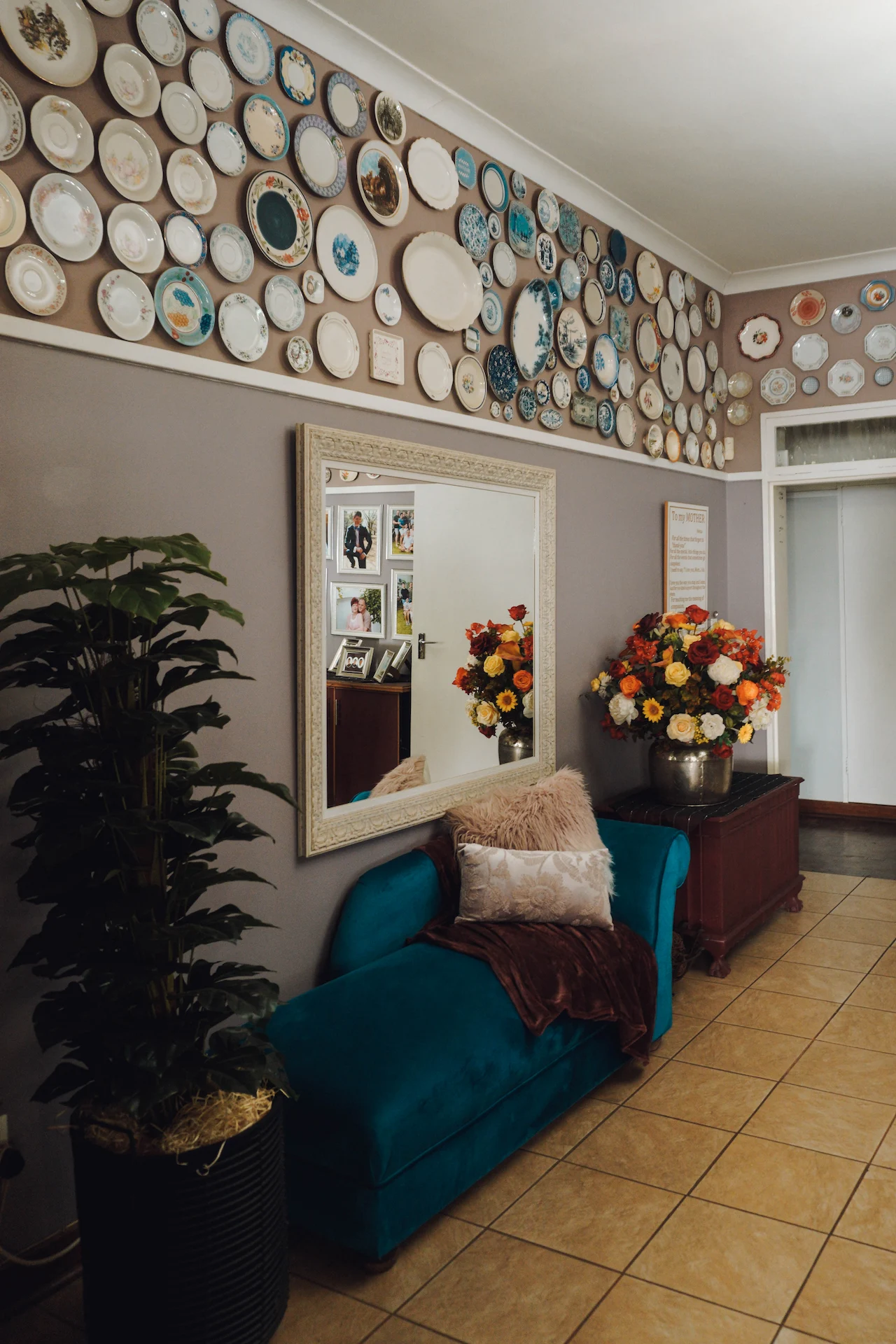 St. Joseph's Guesthouse foyer with a teal chaise lounge, pillows, large mirror, wall plates, flowers, and potted plant.