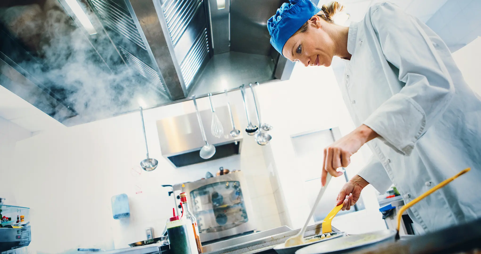 Cuisinière en veste blanche s'affairant sous une hotte d'extraction en pleine vapeur dans une cuisine professionnelle.