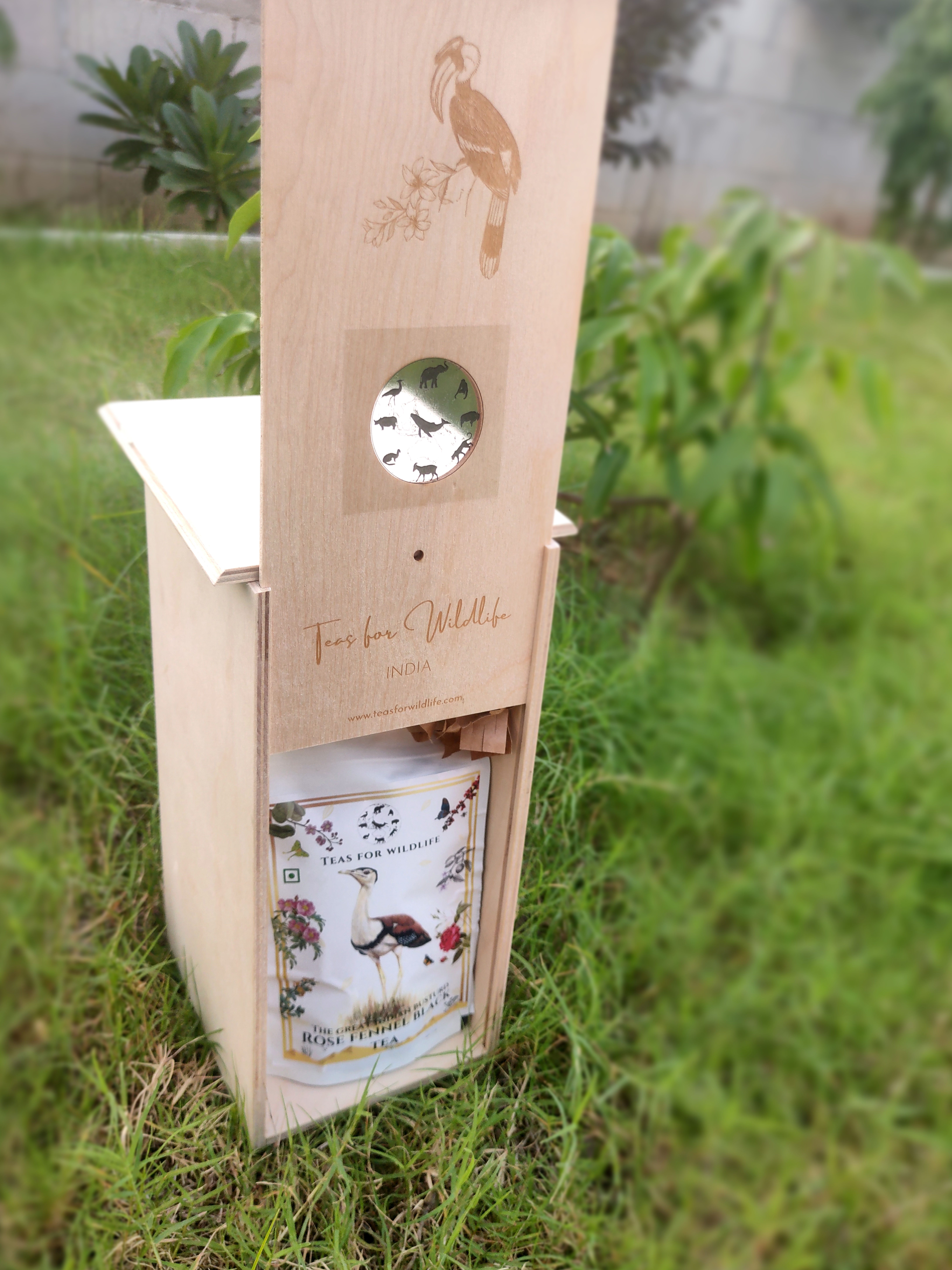 Wooden birdhouse gift box standing on grass outdoors, engraved hornbill on the lid and a tea pouch in its open base.