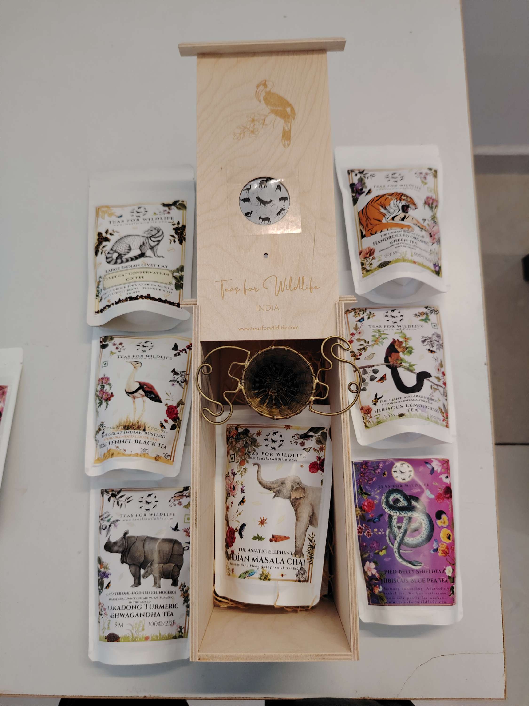 Open wooden birdhouse box viewed from above with a brass strainer inside, ringed by illustrated Teas for Wildlife pouches.